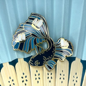 May include: A gold-tone enamel pin featuring a black fish with blue and white fins. The fish is swimming through a wave with three white sailboats.
