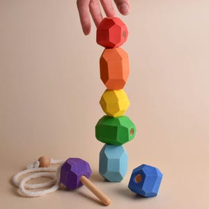 May include: A wooden stacking toy in a rainbow of colours. The stack includes red, orange, yellow, green, and blue geometric shapes. A purple mallet and a blue geometric shape are also present.