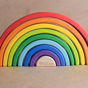 May include: A brightly coloured wooden rainbow stacking toy. The arches are red, orange, yellow, green, blue, and purple. The base has the name "William" engraved. A colourful educational toy for children.