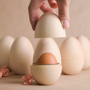 Könnte beinhalten: Ein holzförmiger Behälter in Eiform mit einem braunen Ei im Inneren, wobei eine Hand die Oberseite anhebt. Im Hintergrund befinden sich mehrere andere Holzeier. Der Behälter ist hellbeige und der Hintergrund ist neutral.