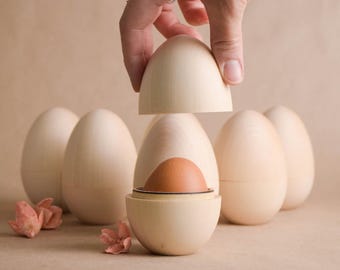 May include: A wooden egg-shaped container with a brown egg inside, with a hand lifting the top. Several other wooden eggs are in the background. The container is light beige, and the background is a neutral tone.