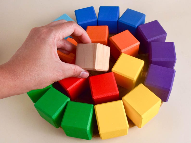 May include: A rainbow-colored set of wooden blocks arranged in a spiral pattern. The blocks are all the same size and shape, and they are made of natural wood. A hand is reaching into the center of the spiral, holding a plain wooden block.