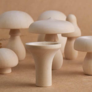 May include: A collection of light-colored wooden mushroom figurines of varying sizes and shapes. Some have rounded caps, while others have flat or slightly pointed tops. The mushrooms are arranged on a neutral-toned surface.