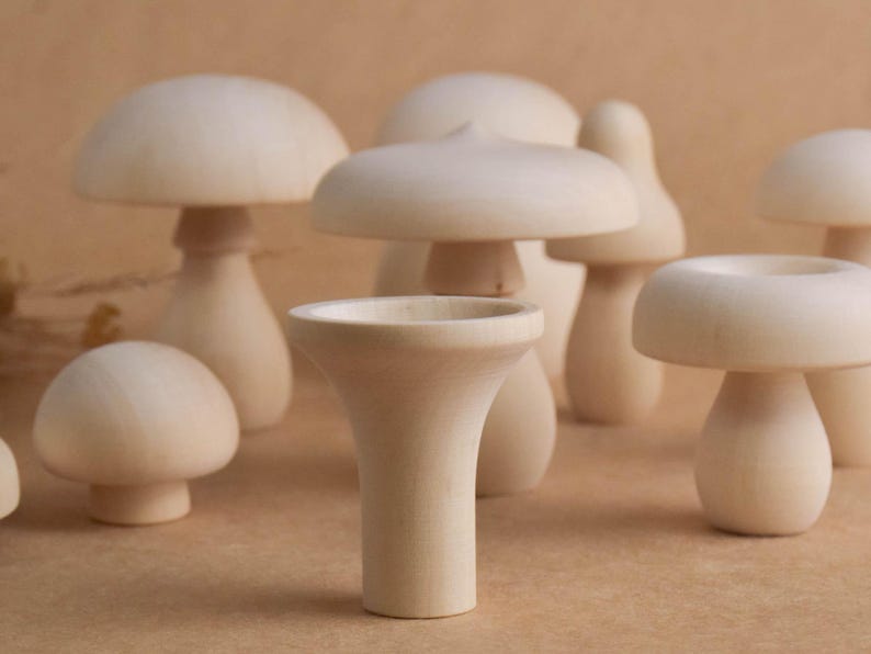 May include: A collection of light-colored wooden mushroom figurines of varying sizes and shapes. Some have rounded caps, while others have flat or slightly pointed tops. The mushrooms are arranged on a neutral-toned surface.