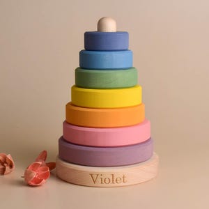 May include: A wooden stacking toy in a rainbow of colours, featuring blue, green, yellow, orange, pink, and purple rings. The base is engraved with the name "Violet". The top has a small, rounded wooden piece.