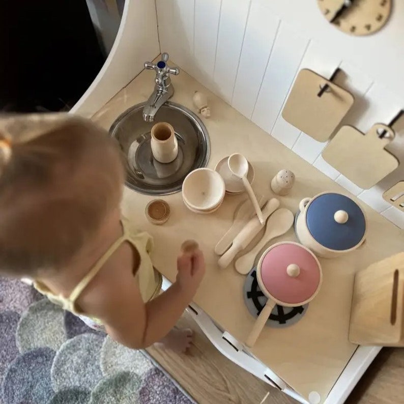 May include: A child plays with a wooden toy kitchen set. The set includes a sink, a stovetop, and various pots, pans, and utensils. The child is wearing a yellow shirt and is standing in front of the kitchen set.