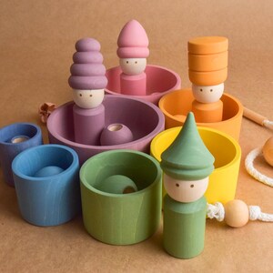 May include: A selection of brightly coloured wooden toys. There are figures with stacked hats and cylindrical cups in shades of pink, purple, orange, yellow, blue, and green. A wooden bead and string are also present.
