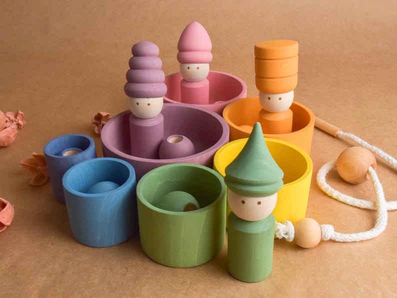 May include: A selection of brightly coloured wooden toys. There are figures with stacked hats and cylindrical cups in shades of pink, purple, orange, yellow, blue, and green. A wooden bead and string are also present.