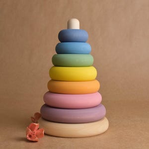 May include: A wooden stacking toy with a natural wood base and a series of colorful rings in shades of blue, green, yellow, orange, pink, and purple. The top piece is a small, white cylinder. The toy is designed for young children.