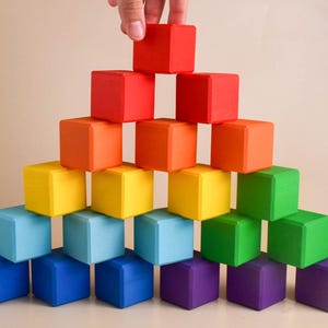 May include: A rainbow-colored wooden block tower with a hand placing the top block. The blocks are arranged in a pyramid shape, with the largest block at the bottom and the smallest block at the top.