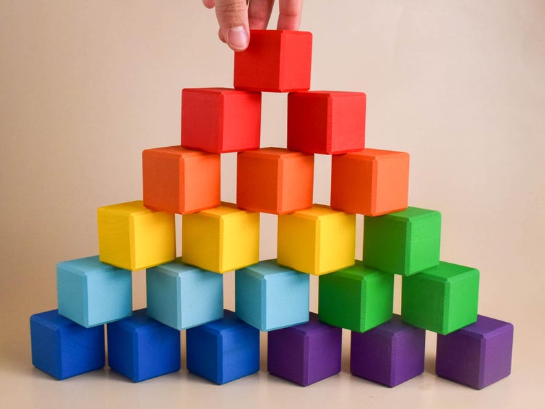 May include: A rainbow-colored wooden block tower with a hand placing the top block. The blocks are arranged in a pyramid shape, with the largest block at the bottom and the smallest block at the top.
