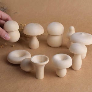 May include: A collection of unfinished wooden mushroom figurines in various shapes and sizes. The mushrooms are light in color and have smooth surfaces. Some have stems, while others are bowl-shaped. The figurines are arranged on a brown surface.