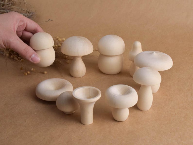 May include: A collection of unfinished wooden mushroom figurines in various shapes and sizes. The mushrooms are light in color and have smooth surfaces. Some have stems, while others are bowl-shaped. The figurines are arranged on a brown surface.