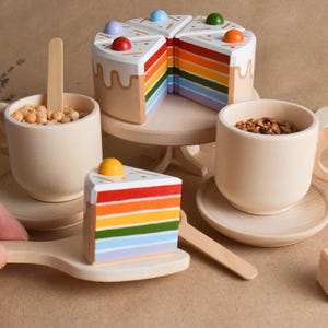 Peut inclure: Un ensemble de thé en bois pour enfants avec un gâteau à étages arc-en-ciel, deux tasses, des soucoupes et une cuillère. Le gâteau est décoré de vermicelles colorés et d'un glaçage blanc.