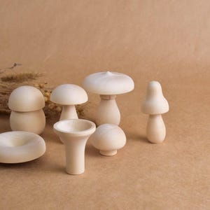 May include: A collection of seven unfinished wooden mushroom figurines in various shapes and sizes. The mushrooms are light beige and sit on a brown surface. Some have rounded caps, while others have more conical shapes. The collection includes a donut-shaped piece.