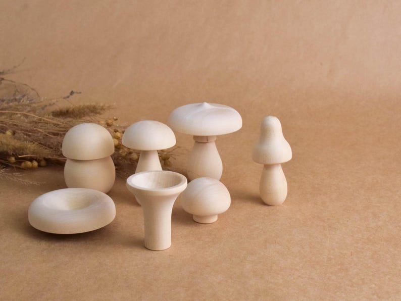 May include: A collection of seven unfinished wooden mushroom figurines in various shapes and sizes. The mushrooms are light beige and sit on a brown surface. Some have rounded caps, while others have more conical shapes. The collection includes a donut-shaped piece.