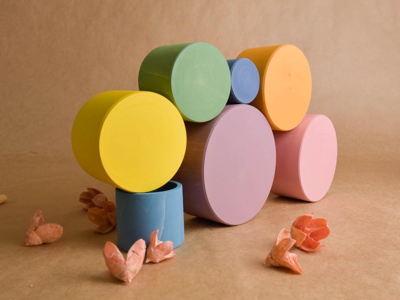 May include: A collection of colourful, cylindrical wooden blocks in shades of yellow, green, blue, orange, purple, and pink. The blocks are stacked, with dried flowers scattered around.