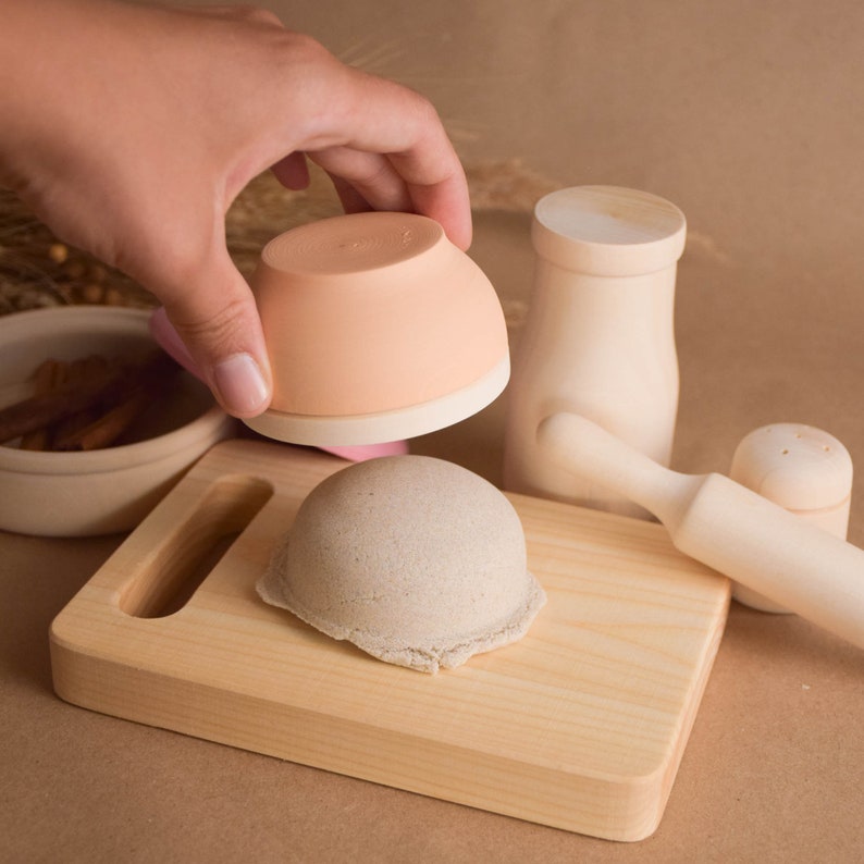 May include: A wooden play kitchen set with a cutting board, rolling pin, salt and pepper shakers, and a small bowl. The bowl is peach colored and the other items are natural wood.