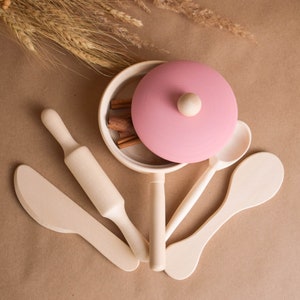 May include: A set of wooden play kitchen utensils, including a rolling pin, a spatula, a spoon, and a pot with a pink lid. The pot has cinnamon sticks inside.