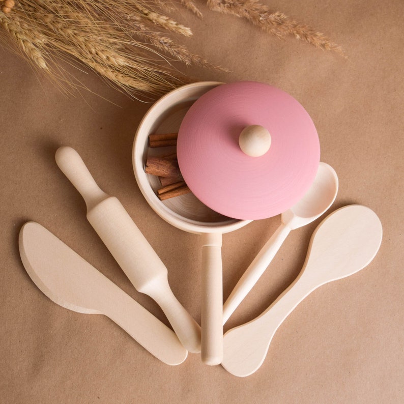 May include: A set of wooden play kitchen utensils, including a rolling pin, a spatula, a spoon, and a pot with a pink lid. The pot has cinnamon sticks inside.