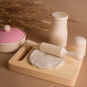 May include: A wooden play kitchen set with a pink lidded container, a rolling pin, a salt shaker, and a milk bottle. The set is on a wooden cutting board with a round piece of play dough.
