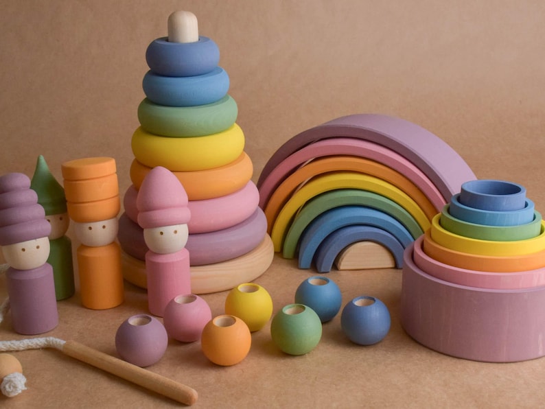 May include: A selection of brightly coloured wooden toys, including a rainbow arch, stacking rings, and peg dolls. The toys are in shades of blue, green, yellow, orange, and pink. The toys are designed for children.