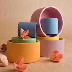 May include: A still life arrangement of colourful, cylindrical ceramic objects in shades of orange, green, blue, pink, and yellow. Dried flower accents are scattered around the arrangement. The background is a neutral tan.