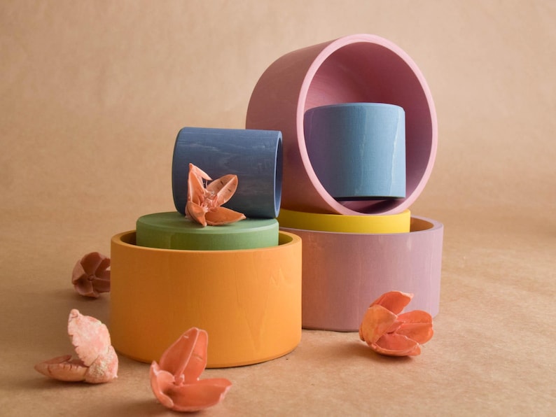 May include: A still life arrangement of colourful, cylindrical ceramic objects in shades of orange, green, blue, pink, and yellow. Dried flower accents are scattered around the arrangement. The background is a neutral tan.