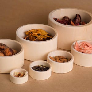 May include: A collection of small, round, off-white wooden bowls filled with various items. The bowls contain nuts, dried fruit, and spices. The bowls are arranged on a light brown surface.