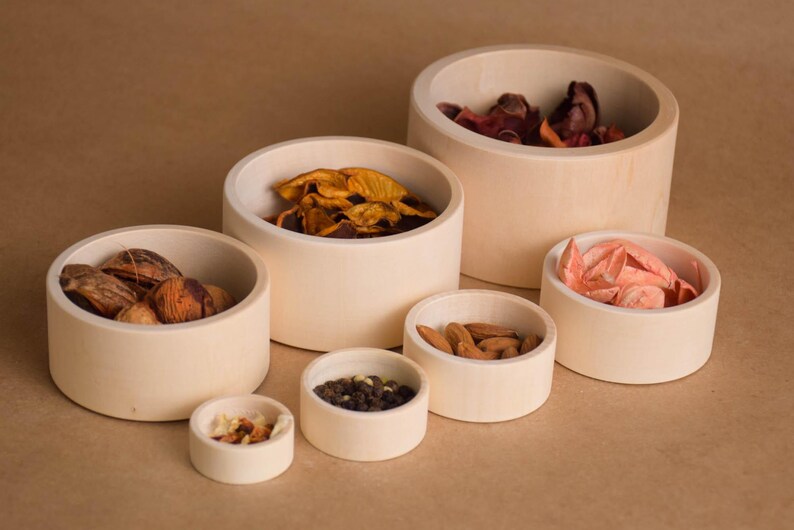 May include: A collection of small, round, off-white wooden bowls filled with various items. The bowls contain nuts, dried fruit, and spices. The bowls are arranged on a light brown surface.