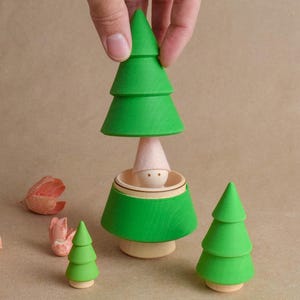 May include: A hand assembling a wooden toy Christmas tree. The toy is green with a light pink gnome inside. Two other green trees and some pink flower petals are also present. The background is a light brown.