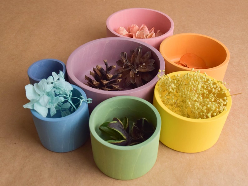 May include: A selection of brightly coloured wooden bowls in different sizes and hues, including pink, orange, yellow, green, blue, and purple. Each bowl contains decorative items such as dried flowers, pine cones, and leaves, creating a vibrant display.