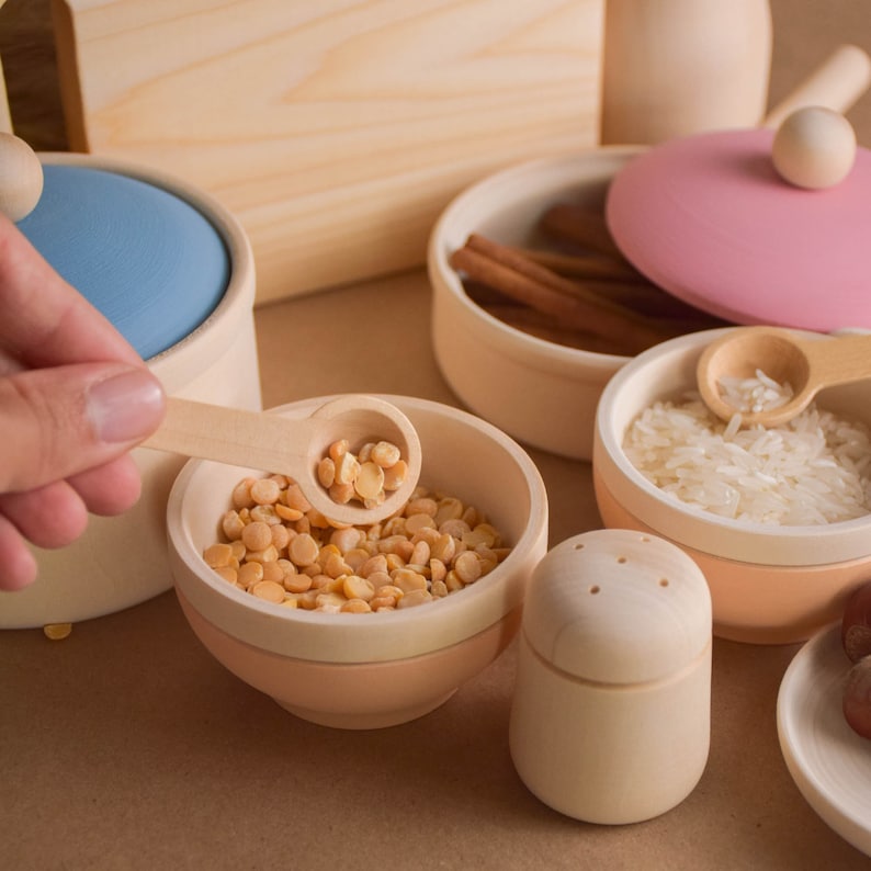 May include: A set of wooden play kitchen accessories, including a small bowl filled with yellow split peas, a bowl with white rice, a wooden salt shaker, and a wooden pot with a pink lid.