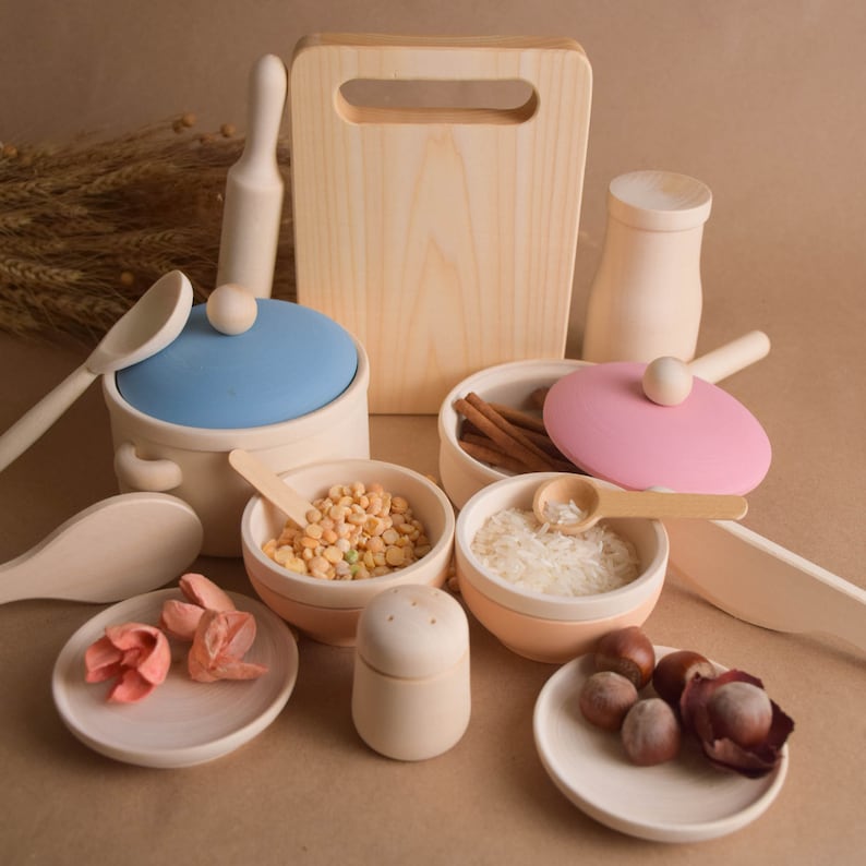May include: A set of wooden play kitchen accessories, including a cutting board, a rolling pin, a pot with a blue lid, a pan with a pink lid, a salt shaker, two bowls, a spoon, and a plate with nuts. The bowls contain dried peas and rice.