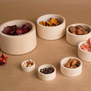 May include: A collection of small, off-white ceramic bowls filled with various items, including dried petals, nuts, and spices. The bowls are arranged on a neutral-toned surface, creating a visually appealing display.
