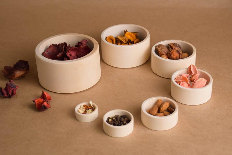 May include: A collection of small, off-white ceramic bowls filled with various items, including dried petals, nuts, and spices. The bowls are arranged on a neutral-toned surface, creating a visually appealing display.