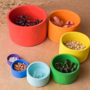 May include: A set of rainbow coloured wooden nesting bowls. The bowls are stacked in a circle with the largest bowl at the top and the smallest bowl at the bottom. Each bowl contains a different item, such as seeds, dried flowers, and small rocks.