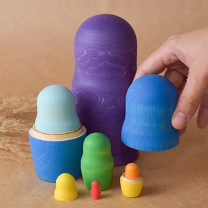 Pode incluir: Um conjunto de bonecas de madeira de encaixe coloridas, tamb&eacute;m conhecidas como bonecas russas. A maior boneca &eacute; roxa, a pr&oacute;xima &eacute; azul, seguida de uma azul claro, verde e uma pequena boneca amarela. As bonecas s&atilde;o pintadas com um padr&atilde;o de gr&atilde;o de madeira.