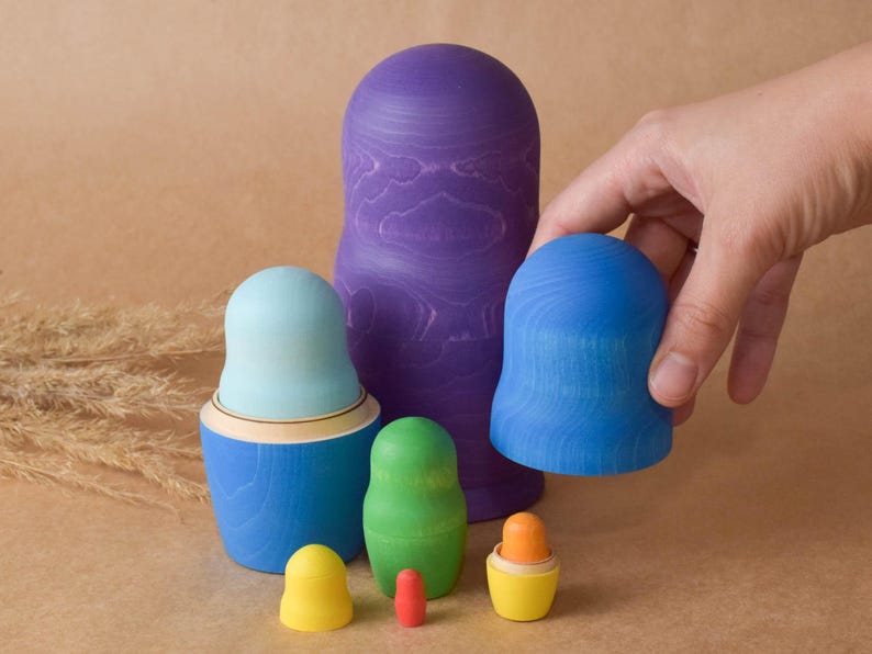 Pode incluir: Um conjunto de bonecas de madeira de encaixe coloridas, tamb&eacute;m conhecidas como bonecas russas. A maior boneca &eacute; roxa, a pr&oacute;xima &eacute; azul, seguida de uma azul claro, verde e uma pequena boneca amarela. As bonecas s&atilde;o pintadas com um padr&atilde;o de gr&atilde;o de madeira.