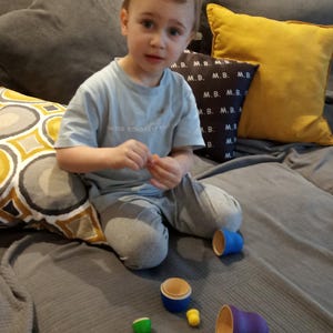 Pode incluir: Uma crian&ccedil;a pequena est&aacute; sentada em um sof&aacute; cinza com almofadas de padr&otilde;es amarelos e cinzentos. A crian&ccedil;a est&aacute; brincando com um conjunto de copos empilh&aacute;veis coloridos. Os copos s&atilde;o de madeira e t&ecirc;m uma variedade de cores, incluindo azul, verde e amarelo.
