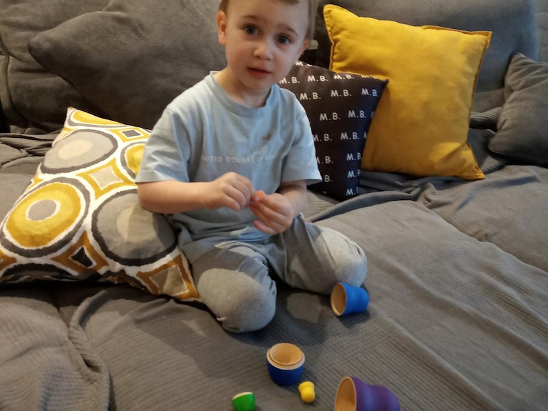 Pode incluir: Uma crian&ccedil;a pequena est&aacute; sentada em um sof&aacute; cinza com almofadas de padr&otilde;es amarelos e cinzentos. A crian&ccedil;a est&aacute; brincando com um conjunto de copos empilh&aacute;veis coloridos. Os copos s&atilde;o de madeira e t&ecirc;m uma variedade de cores, incluindo azul, verde e amarelo.