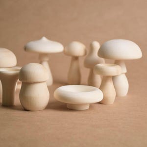 May include: A collection of wooden mushroom figurines in various sizes and shapes. The mushrooms are unpainted, showcasing the natural wood grain. Some have rounded caps, while others have flat or conical tops. The background is a solid, neutral color.