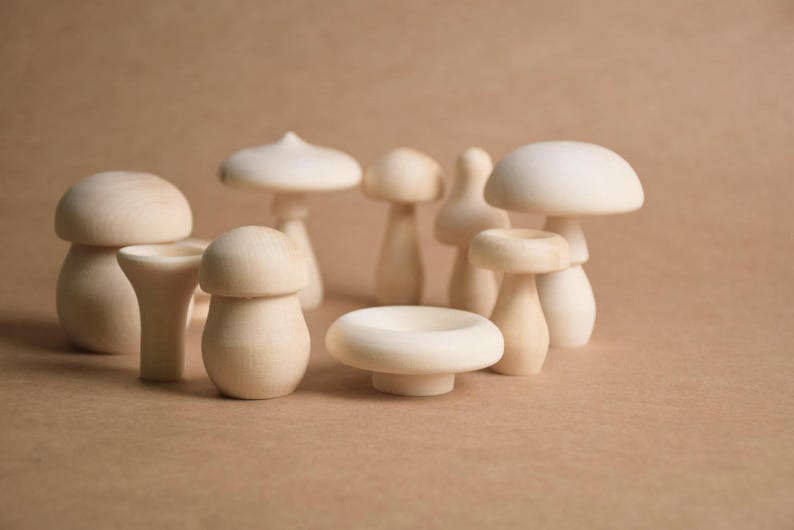 May include: A collection of wooden mushroom figurines in various sizes and shapes. The mushrooms are unpainted, showcasing the natural wood grain. Some have rounded caps, while others have flat or conical tops. The background is a solid, neutral color.
