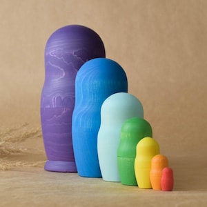 Pode incluir: Um conjunto de bonecas russas de madeira, tamb&eacute;m conhecidas como bonecas de encaixe. As bonecas s&atilde;o pintadas em tons de roxo, azul, verde, amarelo e laranja. A boneca maior &eacute; roxa e a menor &eacute; laranja.