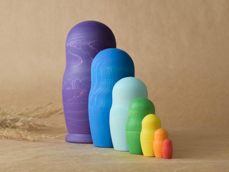 Pode incluir: Um conjunto de bonecas russas de madeira, tamb&eacute;m conhecidas como bonecas de encaixe. As bonecas s&atilde;o pintadas em tons de roxo, azul, verde, amarelo e laranja. A boneca maior &eacute; roxa e a menor &eacute; laranja.