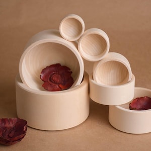 May include: A collection of light-colored wooden bowls of varying sizes, some stacked, with a few containing dark red, textured objects. The bowls are arranged on a neutral-toned surface.