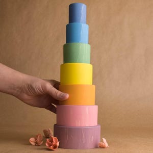 May include: A tower of brightly coloured, cylindrical wooden blocks in shades of blue, green, yellow, orange, pink, and purple. A hand is touching the side of the tower. The blocks are a toy.