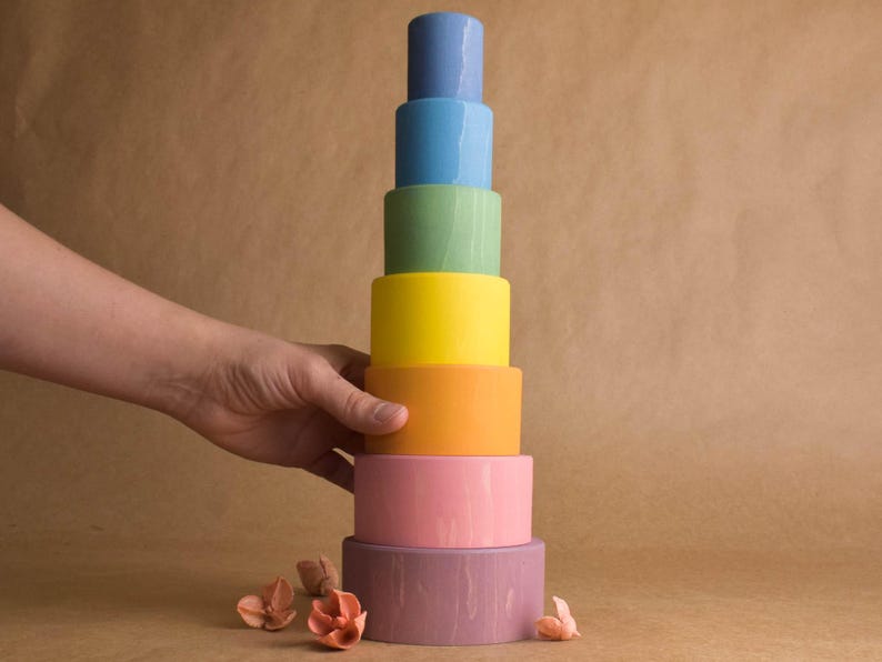 May include: A tower of brightly coloured, cylindrical wooden blocks in shades of blue, green, yellow, orange, pink, and purple. A hand is touching the side of the tower. The blocks are a toy.