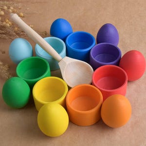 Peut inclure: Un ensemble de jouets en bois colorés, comprenant des bols et des œufs, disposés sur une surface neutre. Une cuillère en bois repose parmi les jouets. Les jouets sont peints dans diverses couleurs, dont le bleu, le vert, le jaune, l'orange, le rouge et le violet.