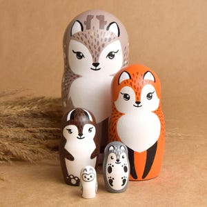 May include: A set of five wooden nesting dolls, each painted with a different woodland animal: a squirrel, fox, hedgehog, owl, and a smaller hedgehog. The dolls are in shades of brown, orange, gray, and white, with black details.